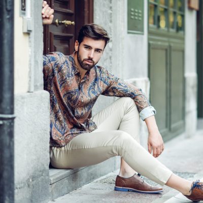 Young bearded man, model of fashion, sitting in an urban step wearing casual clothes. Guy with beard and modern hairstyle looking away in the street.