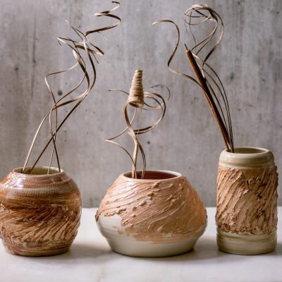 Three different shapes beige brown ceramic vases with dry flowers and twigs branch on white marble table with gray wall behind. Copy space.