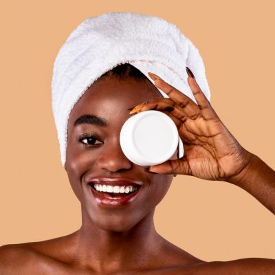Skin Care. Beauty Portrait Of Happy Black Woman Holding White Round Jar With Moisturising Cream In Hand, Covering One Eye With New Skincare Product, Standing Wrapped In Towel Over Studio Background